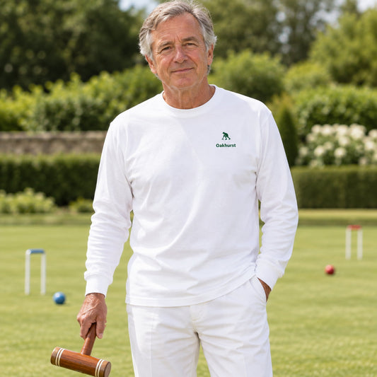 A man standing on a green croquet lawn wearing a white long-sleeve crewneck shirt with an embroidered logo of a croquet striker on the left chest.