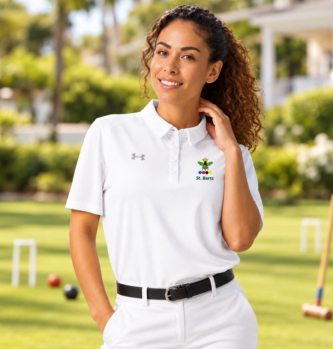 A woman stands on a sunny croquet lawn wearing a white Under Armour polo shirt with a queen bee of croquet embroidered logo on the left chest.