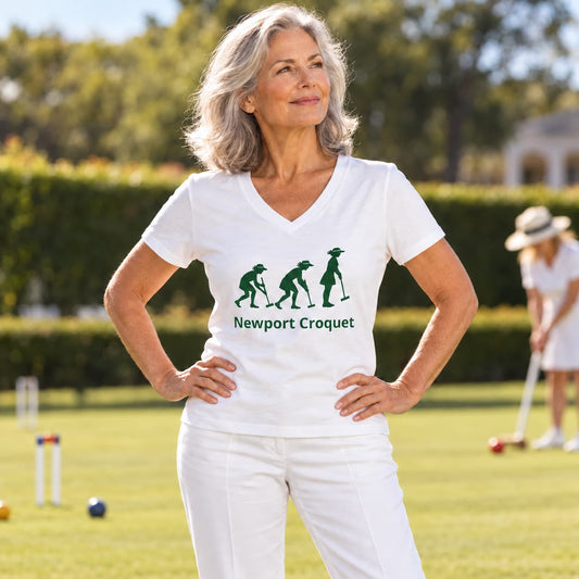 A woman wearing a white V-neck t-shirt with a green croquet graphic of 3 women striking poses on a grassy croquet lawn. The background shows a sunny outdoor garden setting with a blurred figure of another woman playing croquet in the distance.
